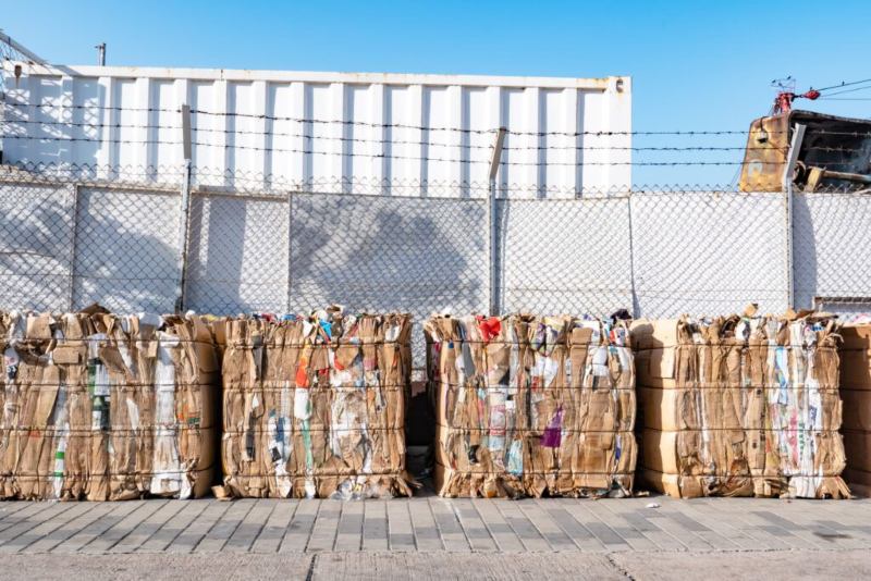Bales of compressed cardboard and recyclables stacked behind a chain-link fence