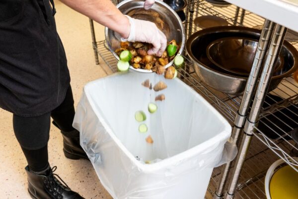 A person dumping food scraps into a trash bin in a commercial kitchen